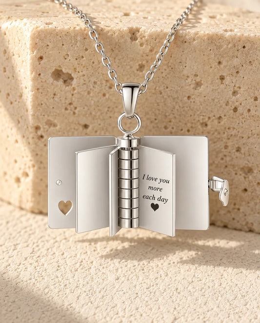 Book locket necklace hanging open, its flat metal pages standing upright around the spine and one page engraved with a love message