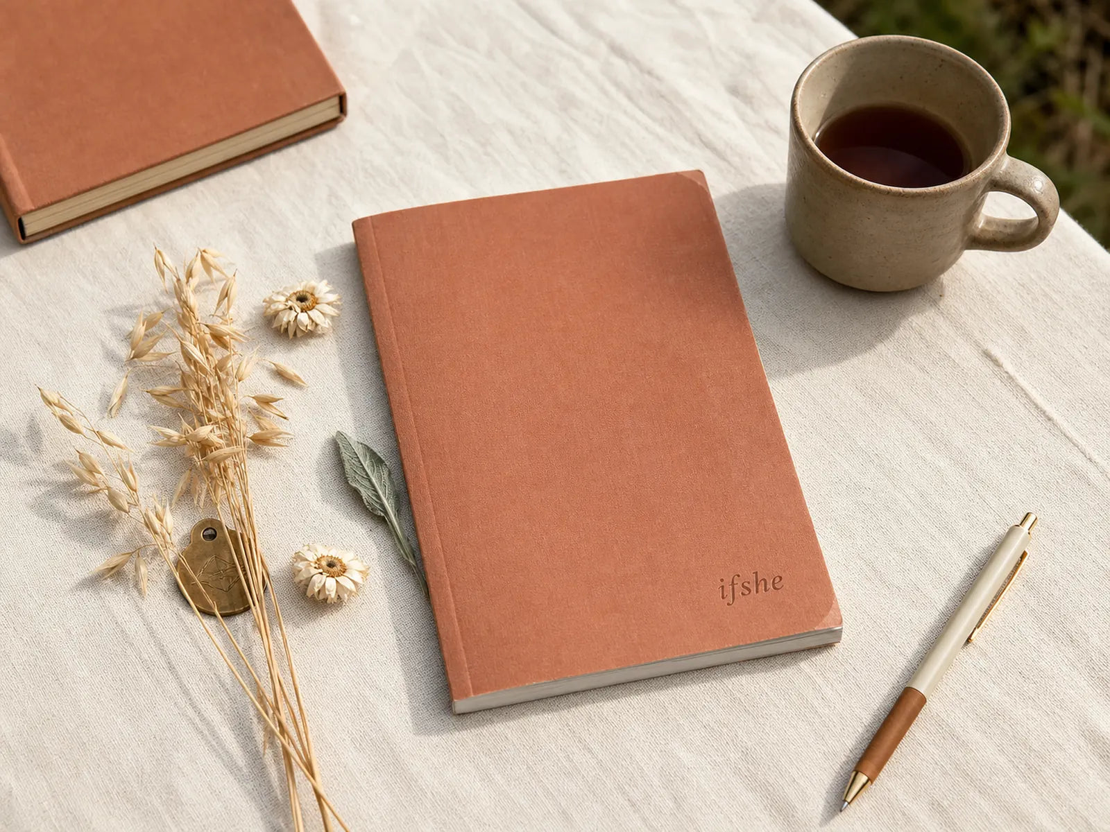 Terracotta cloth-cover pet wedding planning workbook on cream linen with dried wildflowers, brass pet ID tag, ceramic mug of tea, natural outdoor morning light