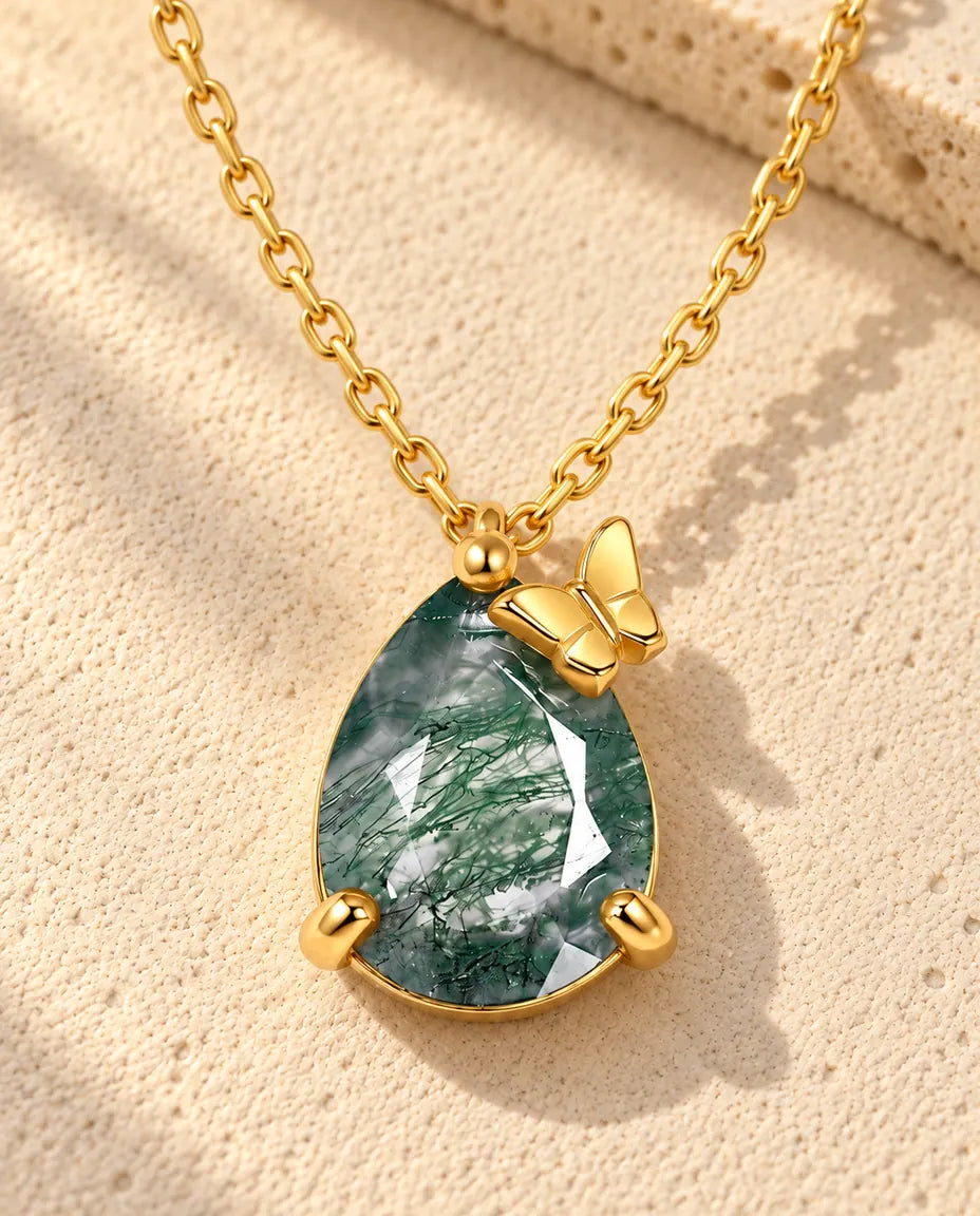 Pear Moss Agate Butterfly Necklace in 10K Gold Plated 925 Sterling Silver - lying on a cream textured surface beside a travertine block