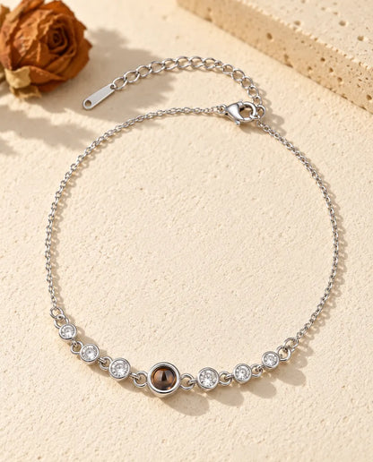 Personalised photo projection bracelet in silver tone with a row of cubic zirconia stones on a fine chain, lying on cream plaster