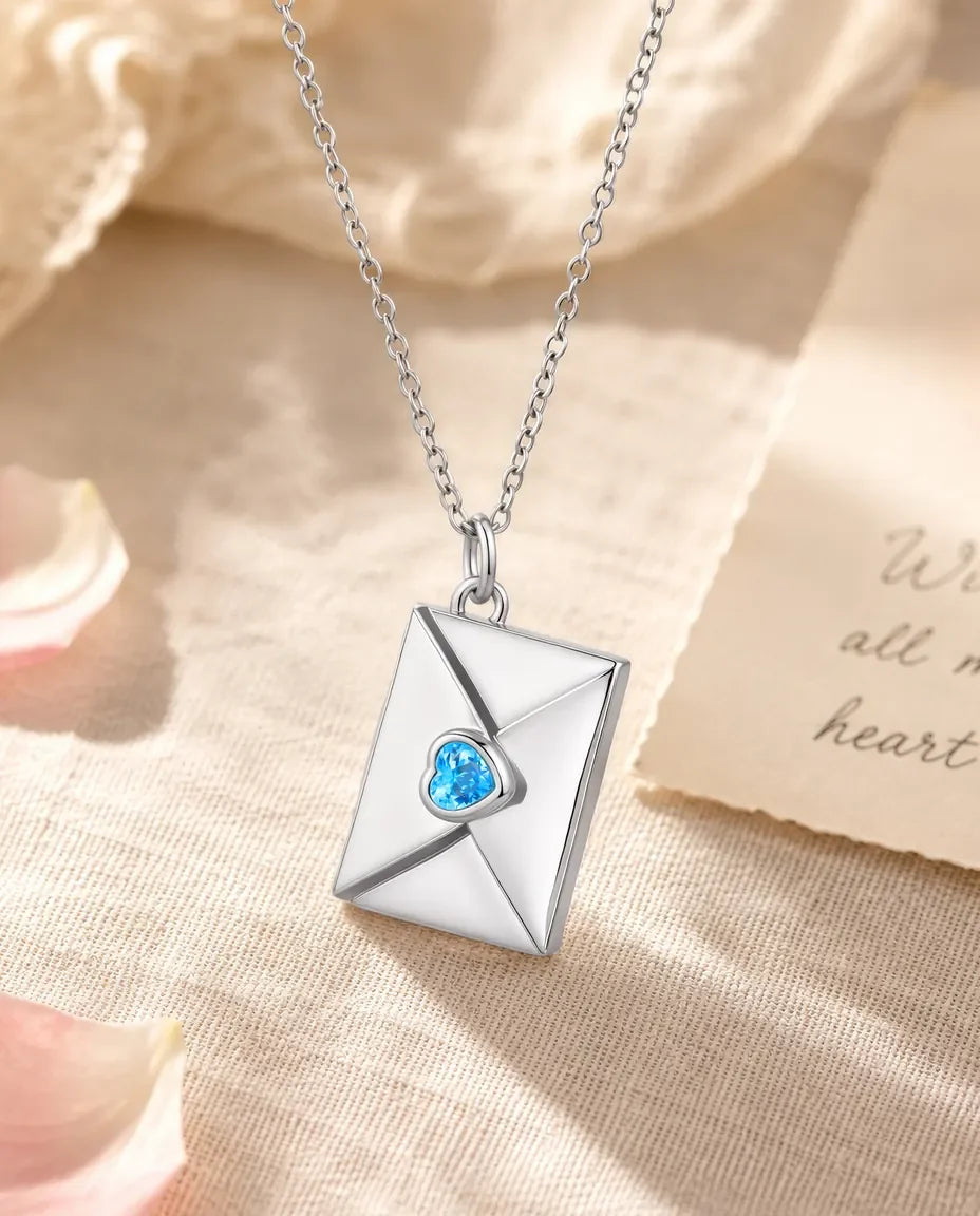 Silver envelope locket necklace with blue heart-cut birthstone on the flap, flat lay