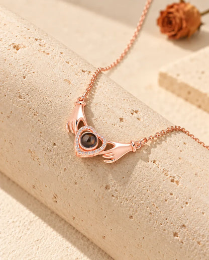 Rose gold tone hands holding heart projection necklace draped over a travertine block