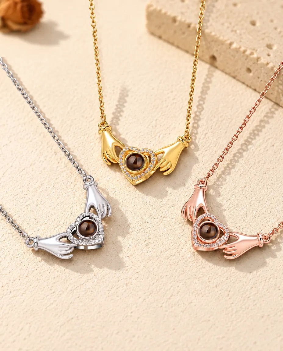 Three colours of the hands holding heart photo projection necklace in a row on cream plaster