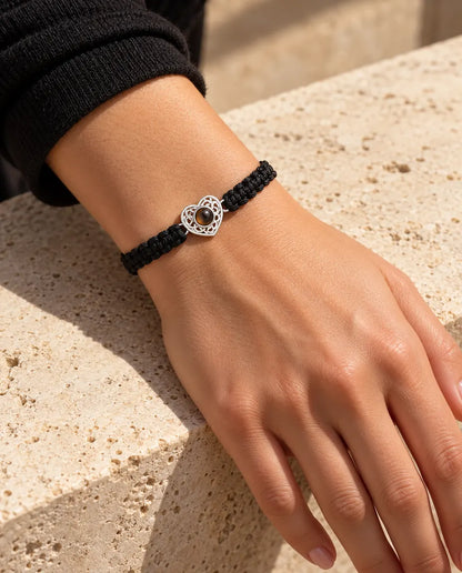 Woman wearing the black braided cord bracelet with a silver tone heart charm on her wrist in warm sunlight