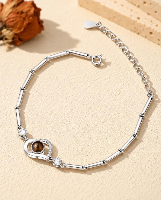 Personalised open heart photo projection bracelet in silver tone on a slim bar chain, lying on cream plaster