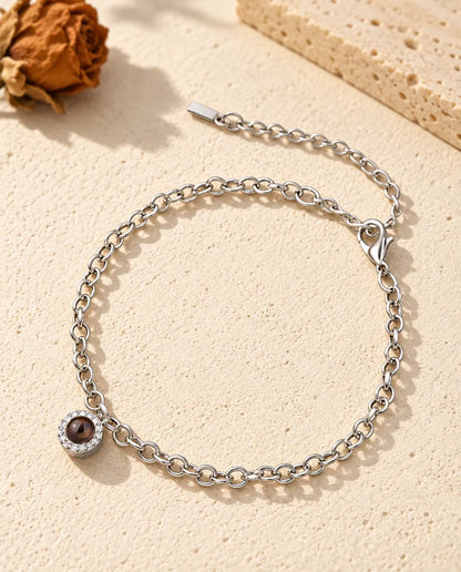 Personalised photo projection bracelet in silver tone with a tiny pave circle charm on a fine chain, lying on cream plaster
