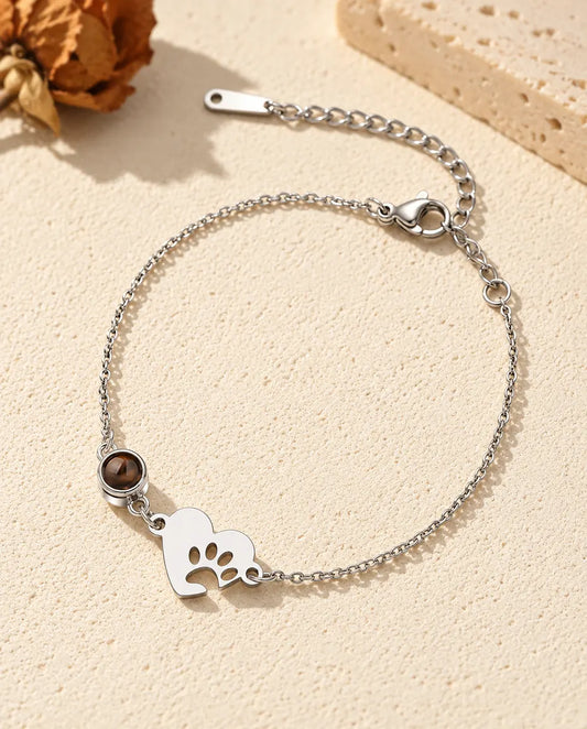 Personalised photo projection bracelet with a silver tone paw print heart tag and projection stone on a fine chain, lying on cream plaster