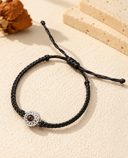 Personalised photo projection bracelet with a silver tone openwork round charm on a black thin braided cord, lying on cream plaster