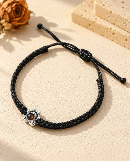 Personalised 925 sterling silver paw charm photo projection bracelet on a black braided cord, lying on cream plaster
